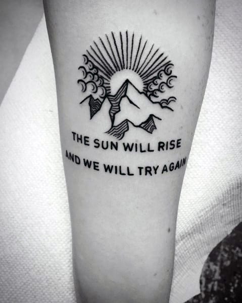 Mountain tattoo on the forearm, featuring bold line peaks, a rising sun with rays, decorative clouds, and an inspiring quote for a motivational design.