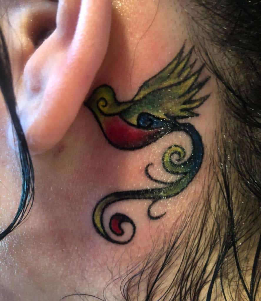 Quetzal tattoo behind ear with swirling tail, featuring shades of green, yellow, red, and blue