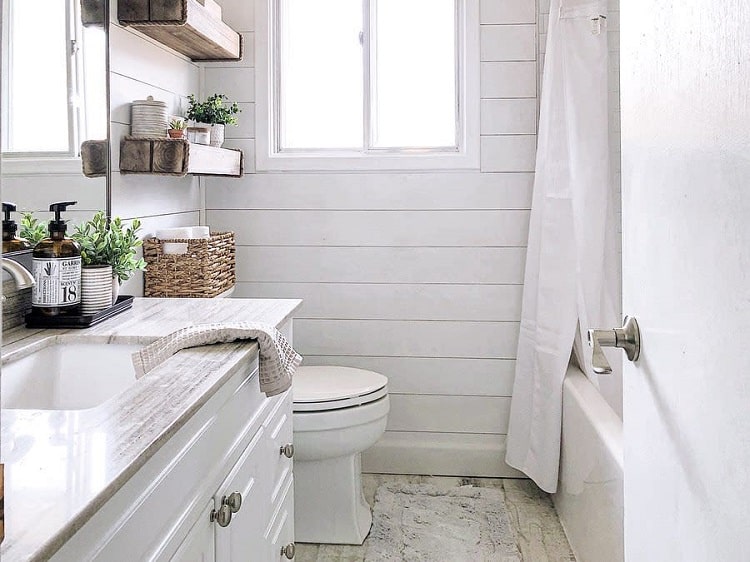 A bright, white farmhouse bathroom with a sink, toilet, and tub, featuring wooden shelves and green plants for decor