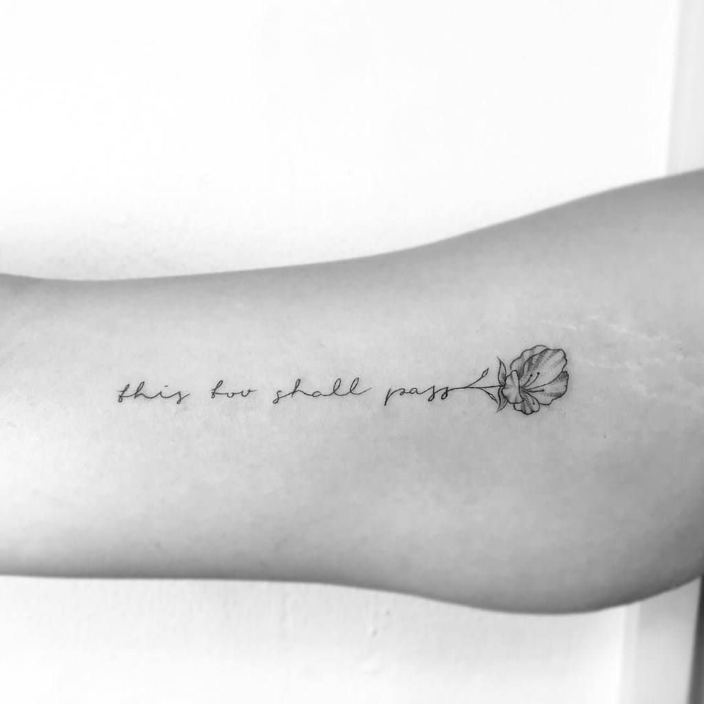 A "This Too Shall Pass" tattoo in cursive with a delicate flower design at the end, on a person's forearm.