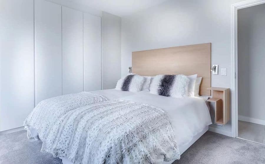 White minimalist bedroom with a large bed, plush pillows, wooden headboard, and wardrobe; soft lighting creates a cozy feel