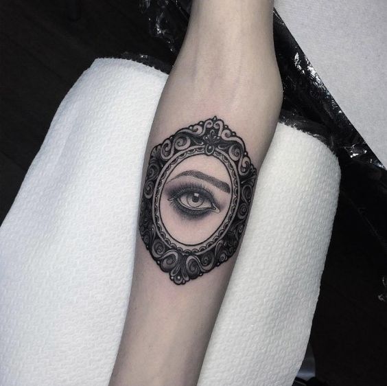 Tattoo of a realistic eye inside an ornate vintage mirror frame, inked in black and grey shading on the forearm.
