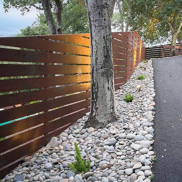 Modern slatted fence with tree, pebble landscaping, and pathway illuminated by soft lighting.