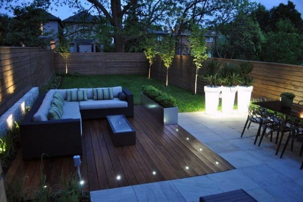 Modern backyard with wooden deck, cozy outdoor seating, ambient lighting, and lush green lawn.