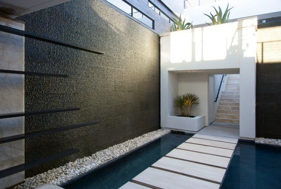 Sleek black wall fountain with a reflective pool and stepping stone walkway in a modern courtyard.