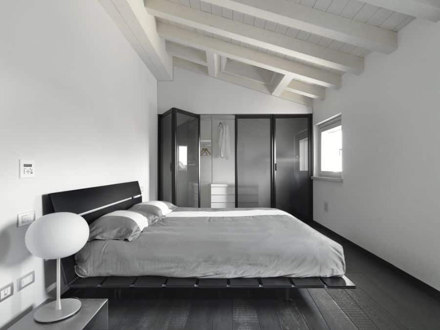 Modern bedroom with black platform bed, gray bedding, exposed beams, and glass wardrobe doors.