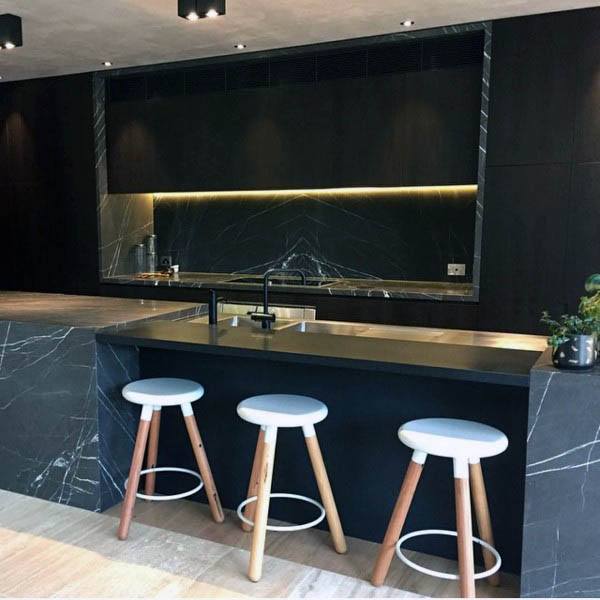 Modern kitchen with black marble countertops, wood stools, and warm under-cabinet lighting.