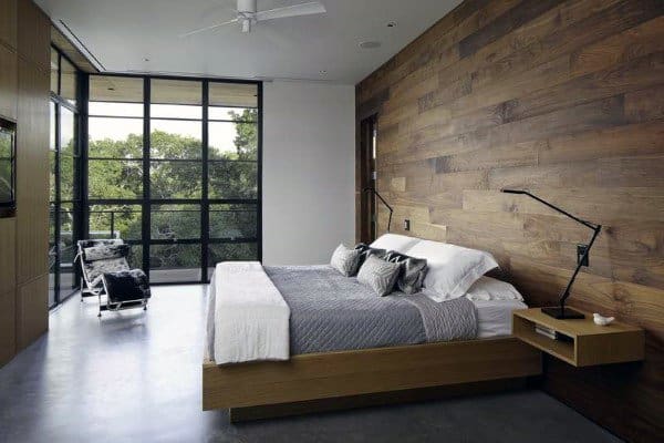 Modern bedroom with wood accent wall, large windows, concrete floors, and a minimalist design featuring a bed and lounge chair