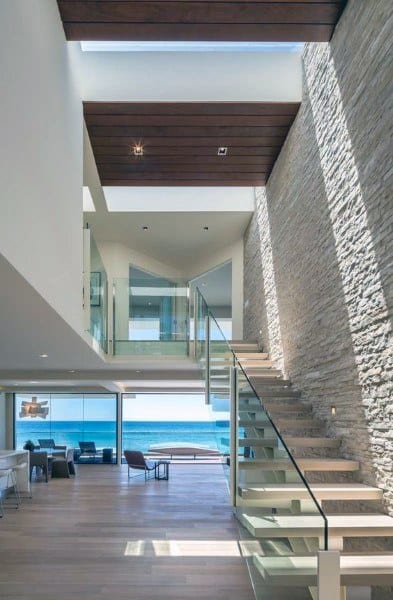 Modern beach house interior with a glass staircase, stone wall, and sweeping ocean views from the living room
