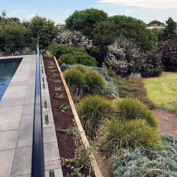A modern poolside patio with lush greenery and a manicured garden in the background under a clear sky