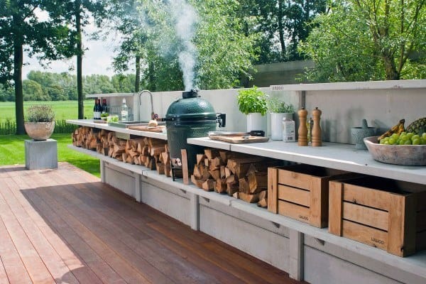 Outdoor kitchen features a green egg smoker, wood storage, and herbs on a wooden deck with a garden view