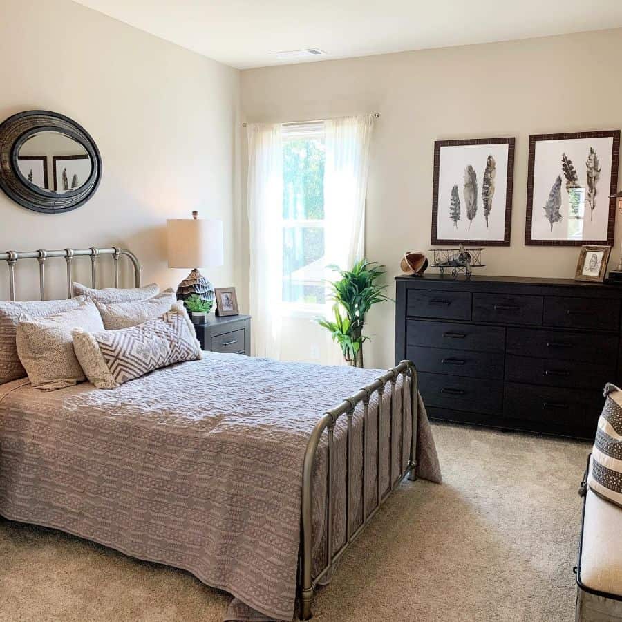 Farmhouse-style cozy bedroom with a metal bed frame, neutral bedding, dresser, round mirror, and framed feather artwork