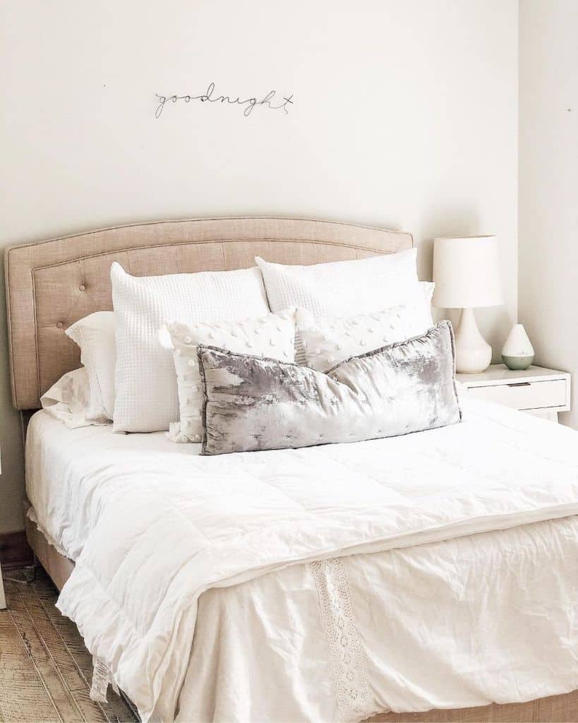 Cozy farmhouse bedroom with a white bed, accent pillows, a beige headboard, and a lamp on a white nightstand