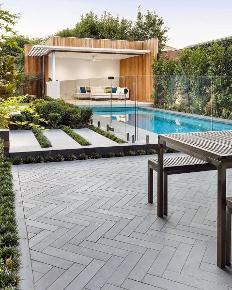 Modern backyard with a pool, glass fence, wooden lounge area, and sleek gray patio