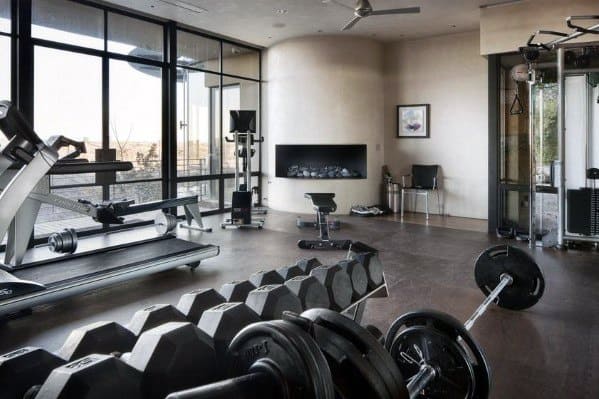 Modern home gym with large windows, treadmill, dumbbells, and a sleek wall-mounted fireplace.