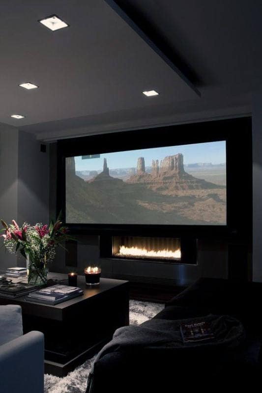 Home theater room with a large screen, cozy lighting, flowers, and a fireplace nestled below the main display