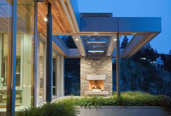 Modern house exterior with large windows, stone fireplace, and surrounding greenery at dusk