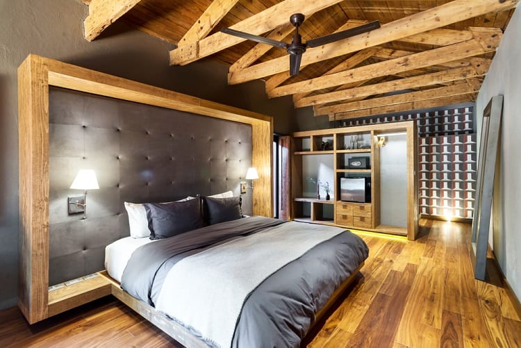 A rustic bedroom with beamed ceilings, a large bed, dark bedding, and modern furniture