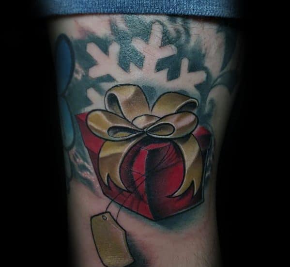 Christmas tattoo of a red gift box wrapped with a golden bow, set against a snowflake background, featuring a blank gift tag for a festive touch.