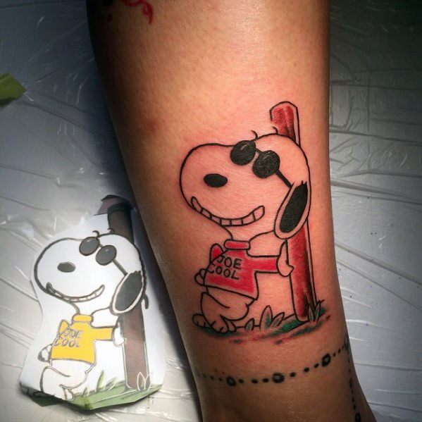 Colorful tattoo of Snoopy as "Joe Cool," wearing sunglasses and a red sweater, leaning against a wooden post, placed on a person's forearm.