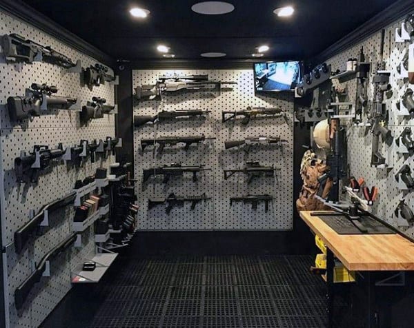 Gun room with pegboard walls displaying firearms, a workbench, and a security monitor.