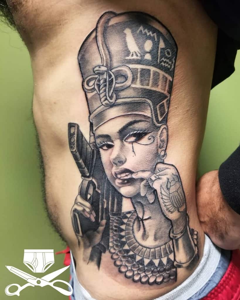 Tattoo of Queen Nefertiti with a gun, adorned in ornate jewelry, on a man's side