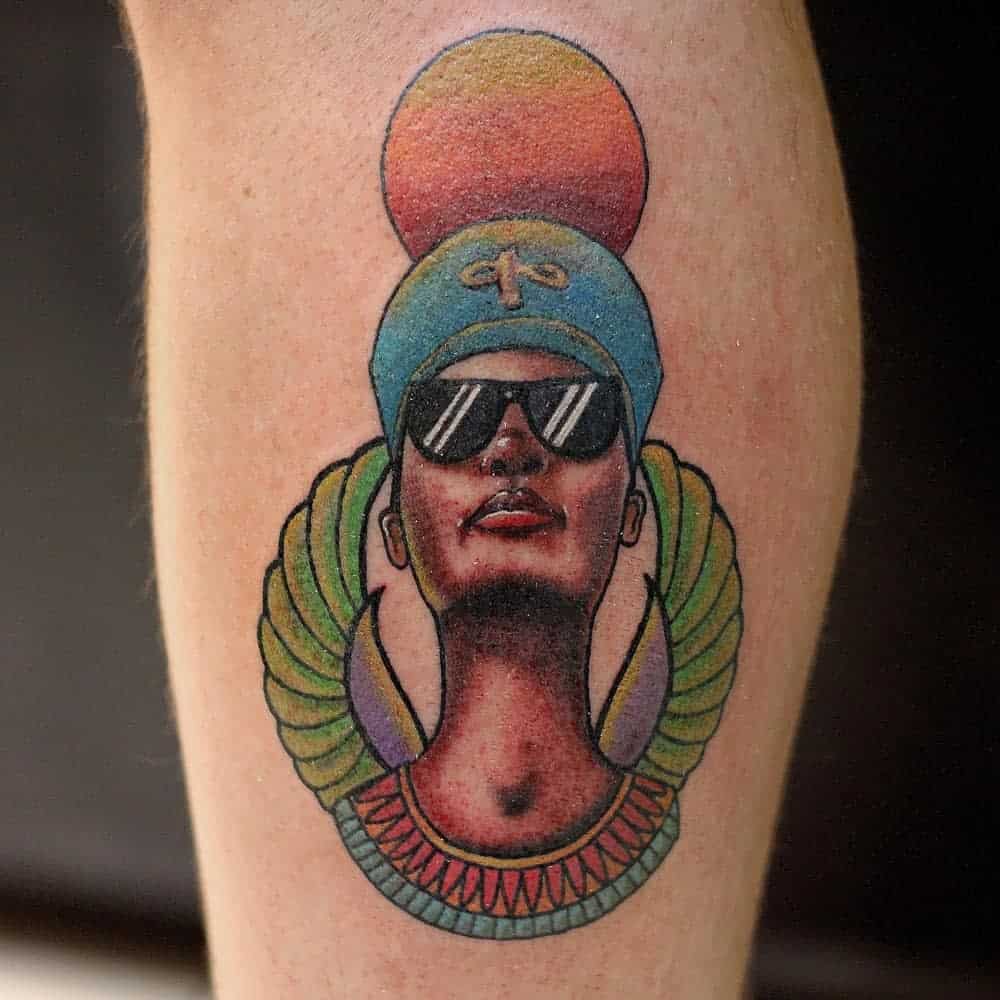 Tattoo of a person with Nefertiti’s headdress, sunglasses, colorful wings, and a sun disk above their head