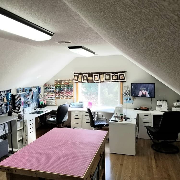 Craft room with organized shelves, sewing machines, and a large pink cutting mat on the table under a sloped ceiling