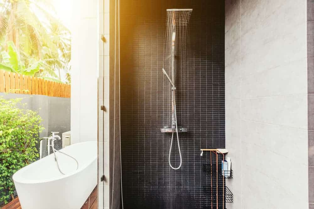 Contemporary outdoor shower with black tiled wall, freestanding bathtub, and lush greenery.