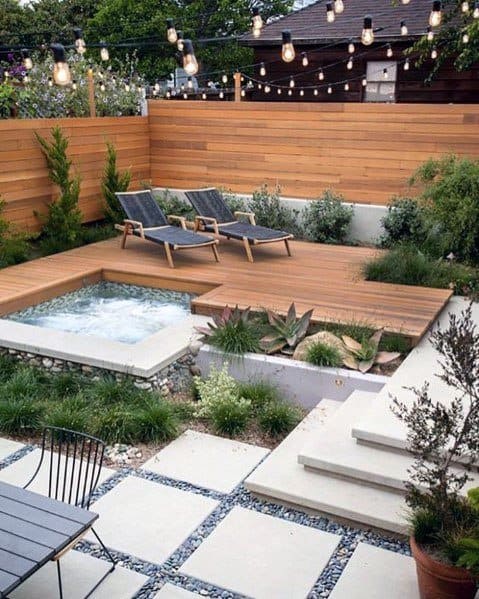 Modern patio design featuring a hot tub, wooden decking, stone steps, and river rock landscaping with lush greenery and stylish lounge chairs