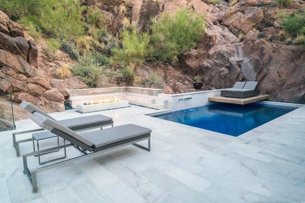 Modern backyard with a sleek pool, fire pit, and minimalist lounge chairs by rocky terrain.