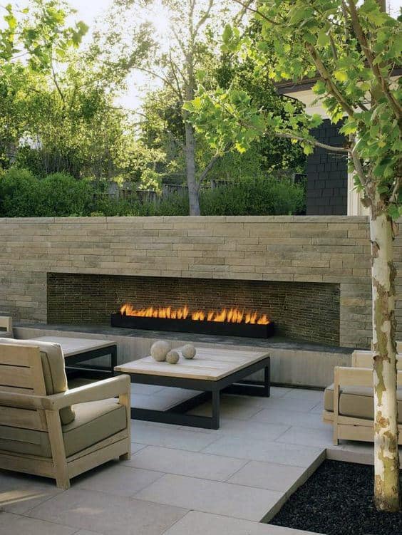 Outdoor patio with a modern fireplace, wooden chairs, and a table, surrounded by greenery and trees