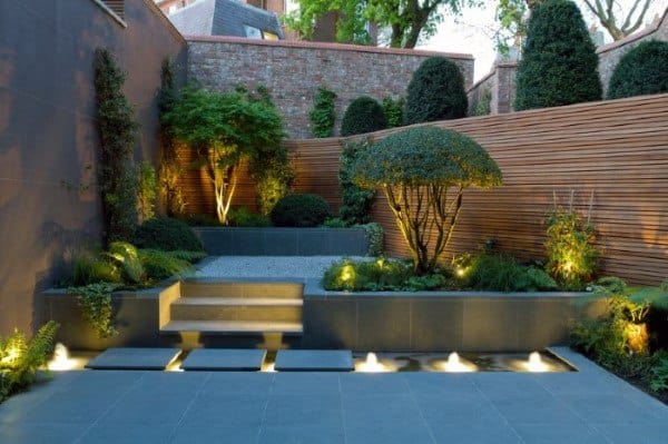 Contemporary courtyard with wooden fencing, lit pathways, trimmed shrubs, and layered greenery.