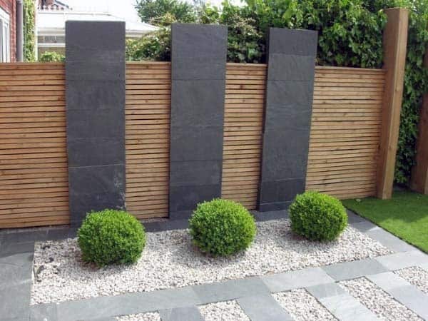 Minimalist garden with gravel, round boxwood shrubs, and vertical slate panels against a wooden fence.