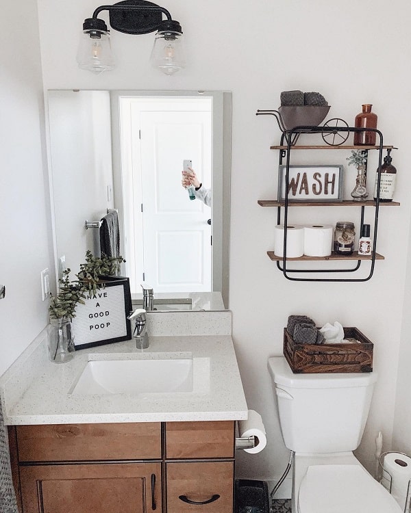 A small, farmhouse-style bathroom with a sink, mirror, and toilet; shelves hold decor; a humorous sign is on the counter