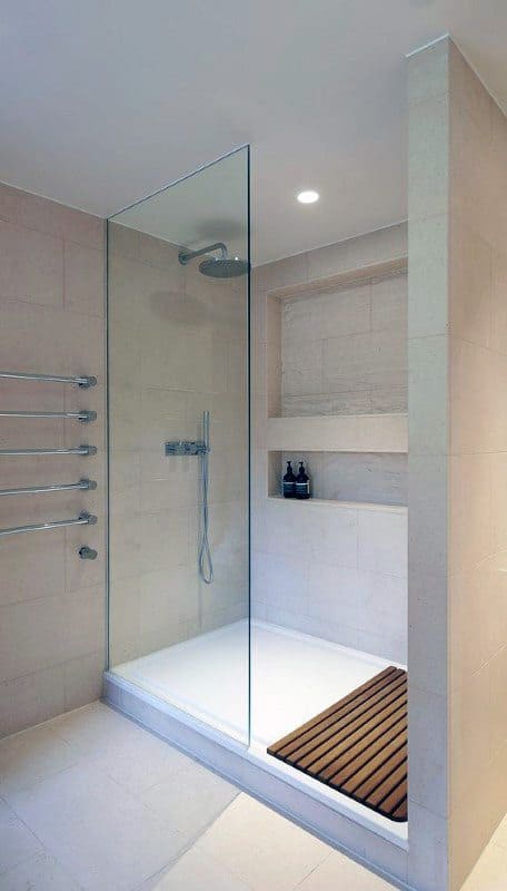 Minimalist shower with glass enclosure, beige tiles, built-in shelves, and wooden floor mat.