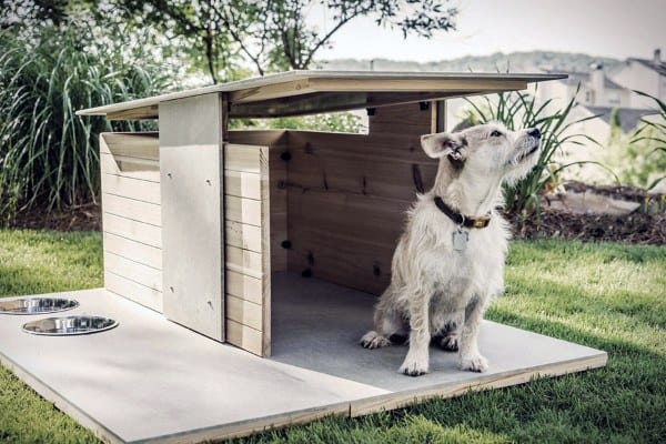 Simple modern dog house with a slanted roof, wooden slats, and two food bowls outside, with a dog standing nearby, set in a grassy area