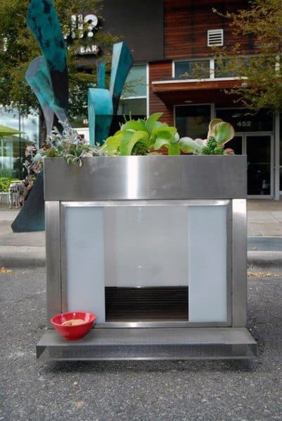 Modern steel dog house with frosted glass panels, a plant-filled top, and a red food bowl outside, located in an urban setting