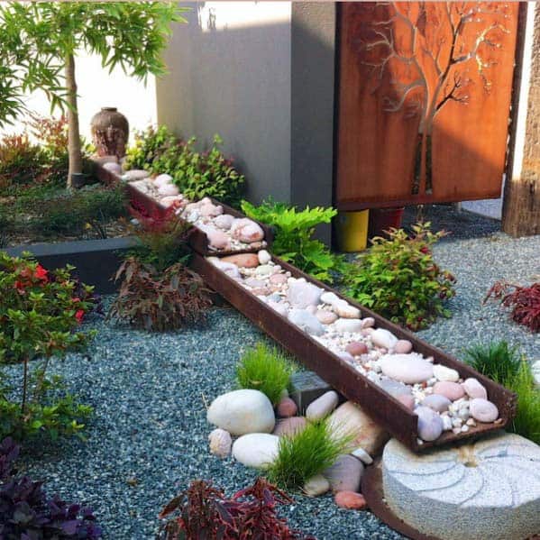 Modern sleek river rock landscaping with a steel channel filled with smooth pebbles, surrounded by vibrant plants and a decorative metal tree panel.