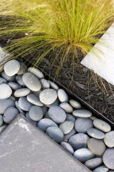 Smooth, rounded stones line a modern garden pathway, bordered with black mulch and complemented by lush greenery