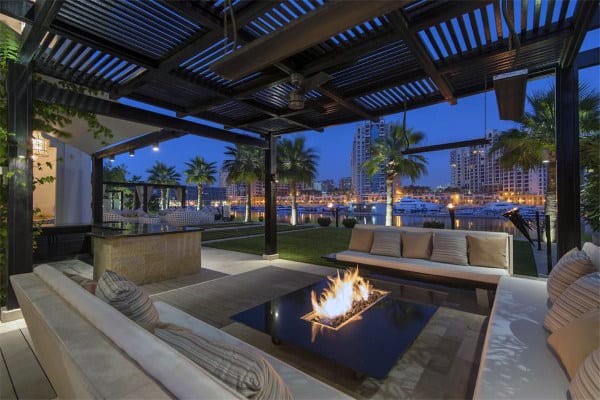 Outdoor lounge with a fire pit, overlooking a marina with palm trees and lit-up city buildings at dusk
