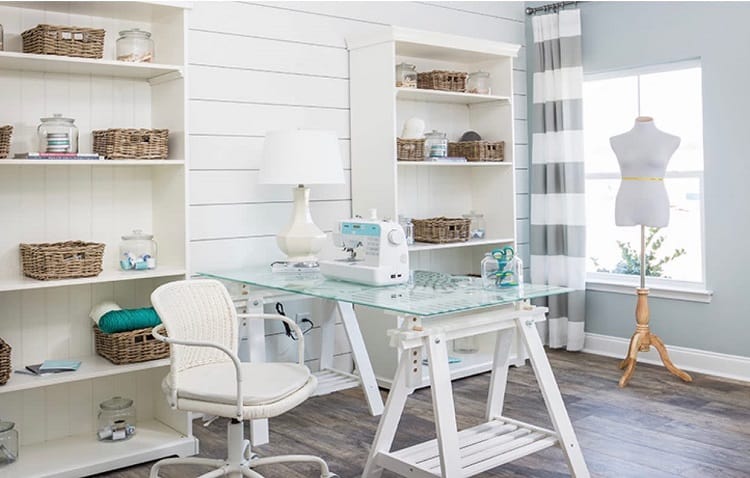 Bright craft room with a sewing machine on a glass desk, wicker baskets on shelves, and a dress form by the window