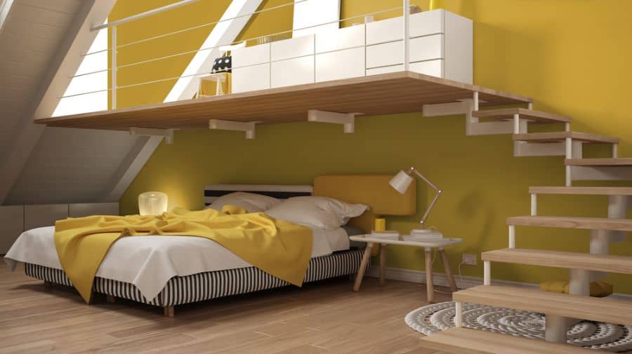Loft bedroom with sunny yellow walls, a bed, bedside table with lamp, wooden stairs, and a small loft area above