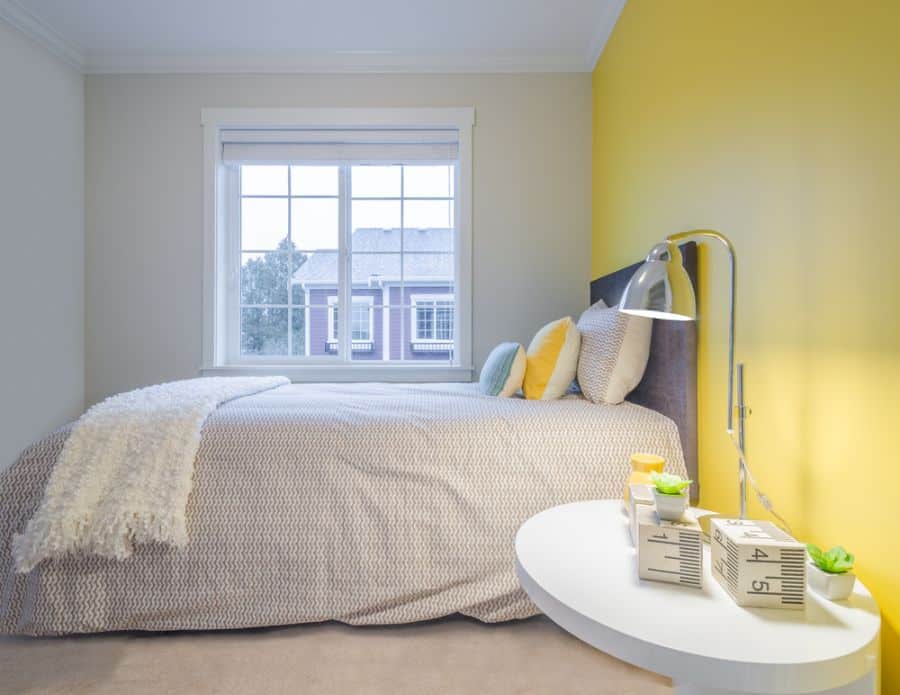 Cozy bedroom with a sunny yellow accent wall, bed by window, and a side table with decor items and a metal lamp