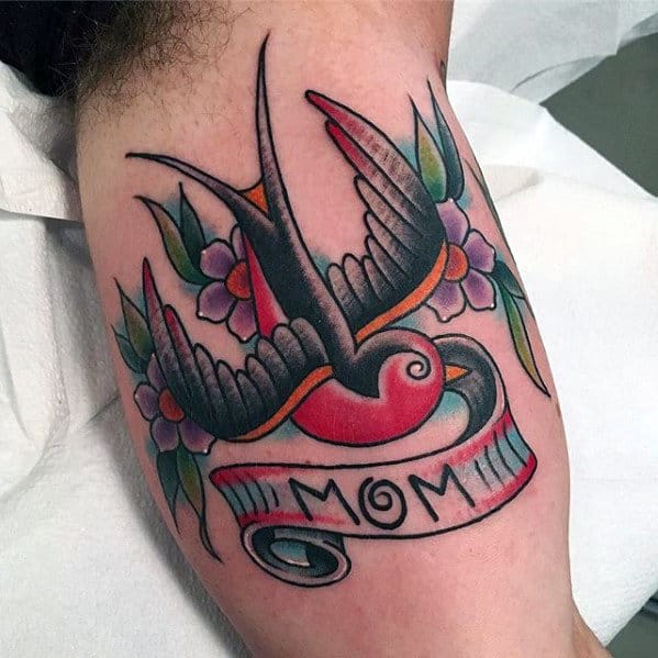 Tattoo of a swallow with pink flowers and a banner reading "MOM" on an arm