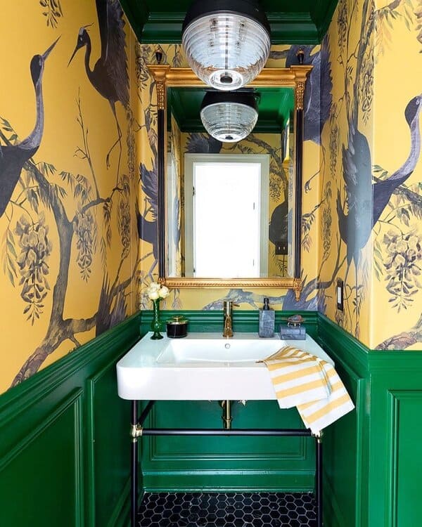 A powder room with crane-printed yellow wallpaper, green wainscoting, gold mirror, and black hex flooring for a dramatic statement.