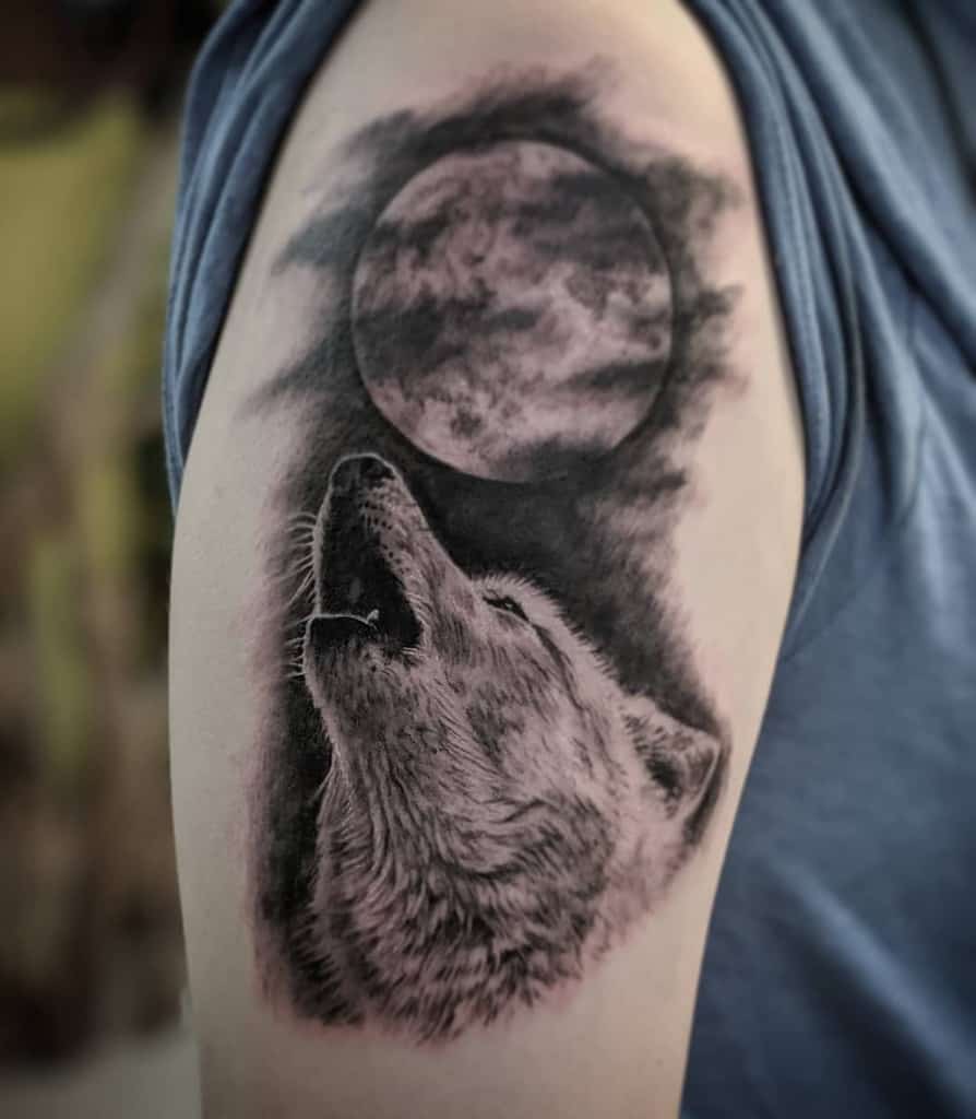 A stunning tattoo of a lone wolf howling beneath a veil of clouds that cloak the moon, beautifully etched on a person's upper arm