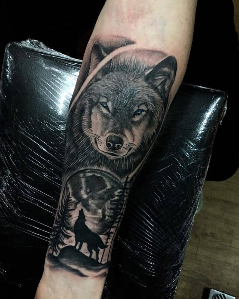 Black and grey forearm tattoo of a wolf's face with piercing blue eyes, accompanied by a silhouette of a howling wolf under a full moon and trees