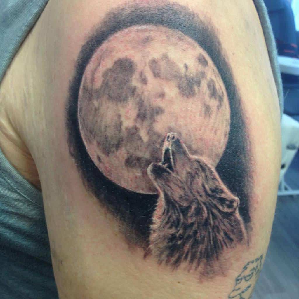 A tattoo of a majestic wolf howling at a luminous full moon graces the person's upper arm