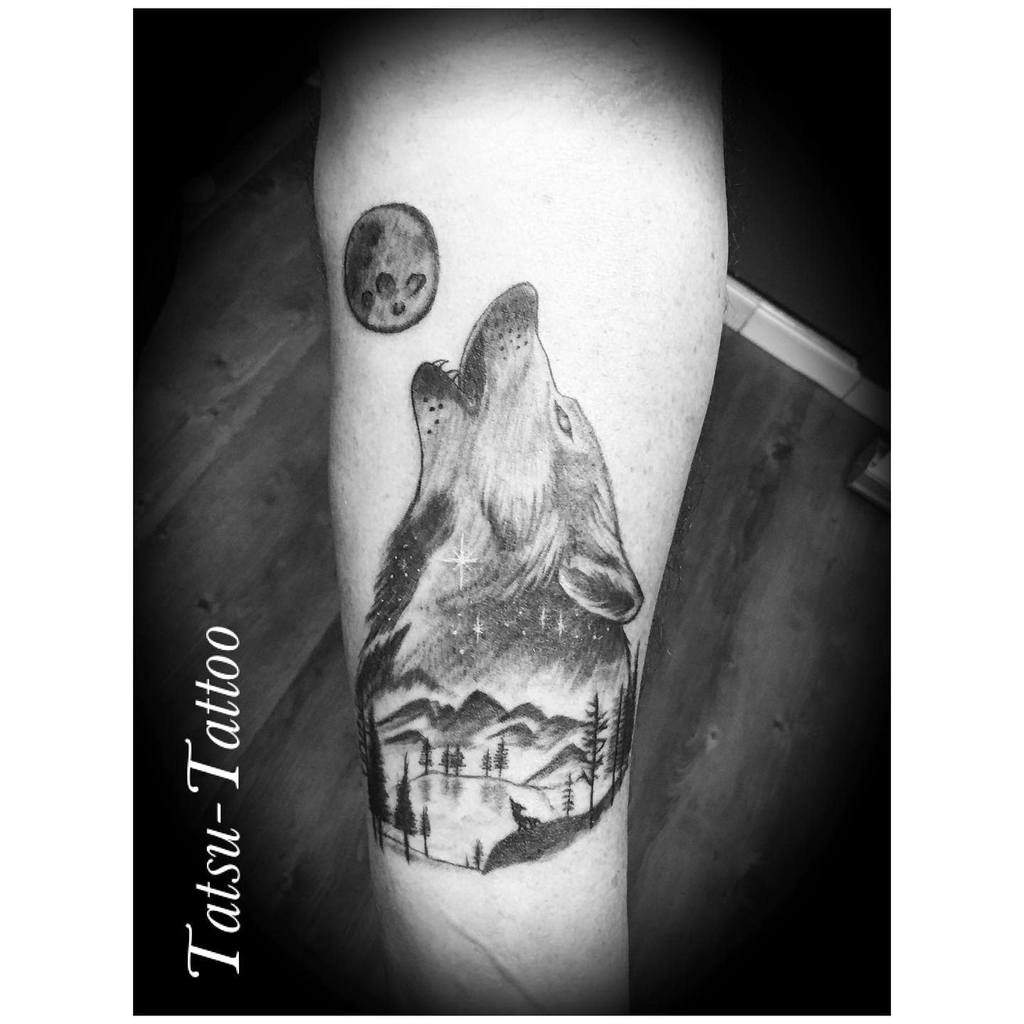 A striking tattoo of a wolf howling, seamlessly blending with a forest landscape and a luminous moon, adorns the person's arm